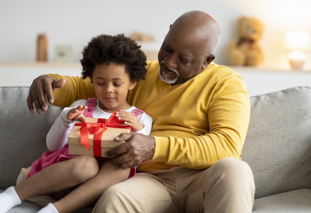 Glad happy cute african american preschool kid and old man open birthday present in living room